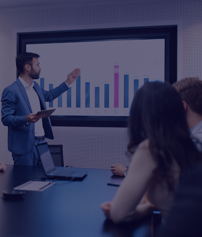 A senior Prodest health insurance agent presents a customized health insurance program to a group of young health insurance agents during a specialized training session for new Prodest agents. A bar chart showcasing deductible options is displayed on a large screen in the conference room, with the young agents facing the presenter and the screen.