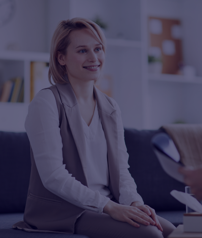 A young woman with shoulder-length hair, wearing a light-colored blazer, sits on a couch in an office setting, smiling at an off-camera health insurance agent while discussing health plans for individuals. The Prodest Insurance offices are visible in the background, lending a professional air to the meeting.