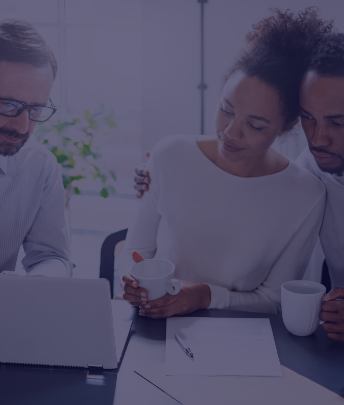 A middle-aged couple sits with a Prodest health insurance advisor and reviews family health insurance plans. The woman holds a mug and has her arm around the man. The advisor, a senior Prodest insurance agent, points at coverage options on his laptop. The couple looks happy and appears to be comfortable with the health insurance options being presented to them.